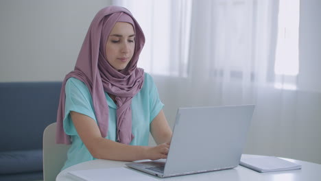 Schöne-Junge-Muslimische-Frau-Arbeitet-Am-Laptop-An-Ihrem-Arbeitsplatz.-Eine-Junge-Muslimische-Frau,-Die-Vor-Einem-Laptop-Bildschirm-Sitzt,-Schaut-Nach-Oben-Und-Blickt-In-Die-Webcam